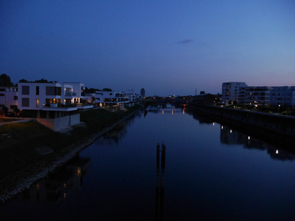 Das Foto ist eine Landschaftsaufnahme, die während der "Blauen Stunde" nach Sonnenuntergang entsanden ist. Daher ist das ganze Bild überproportional Blau. Man sieht ein Hafenbecken, links sind und rechts sind moderne, recht teuer aussehende Häuser. Man sieht wie sich die Lichter im Wasser spiegeln.