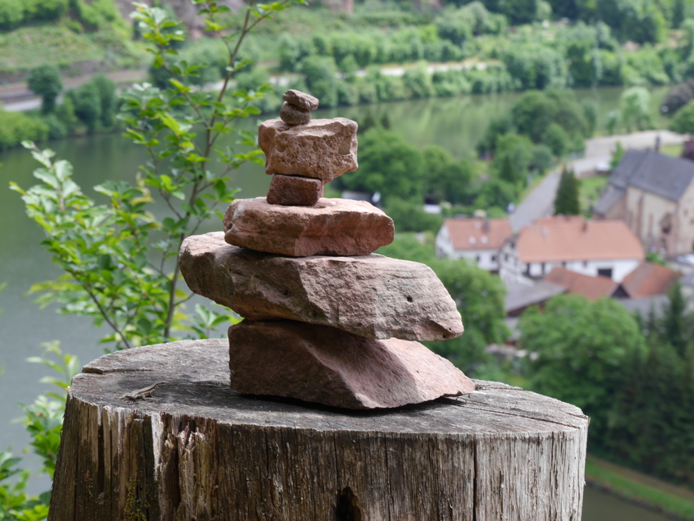 Ein Stapel von kleinen Steinen, der offensichtlich von Kindern auf einem Baumstumpf aufgeschichtet wurde. Dahinter kann man in der Unschärfe des Hintergrunds den Neckar und ein paar Häuser erkennen.