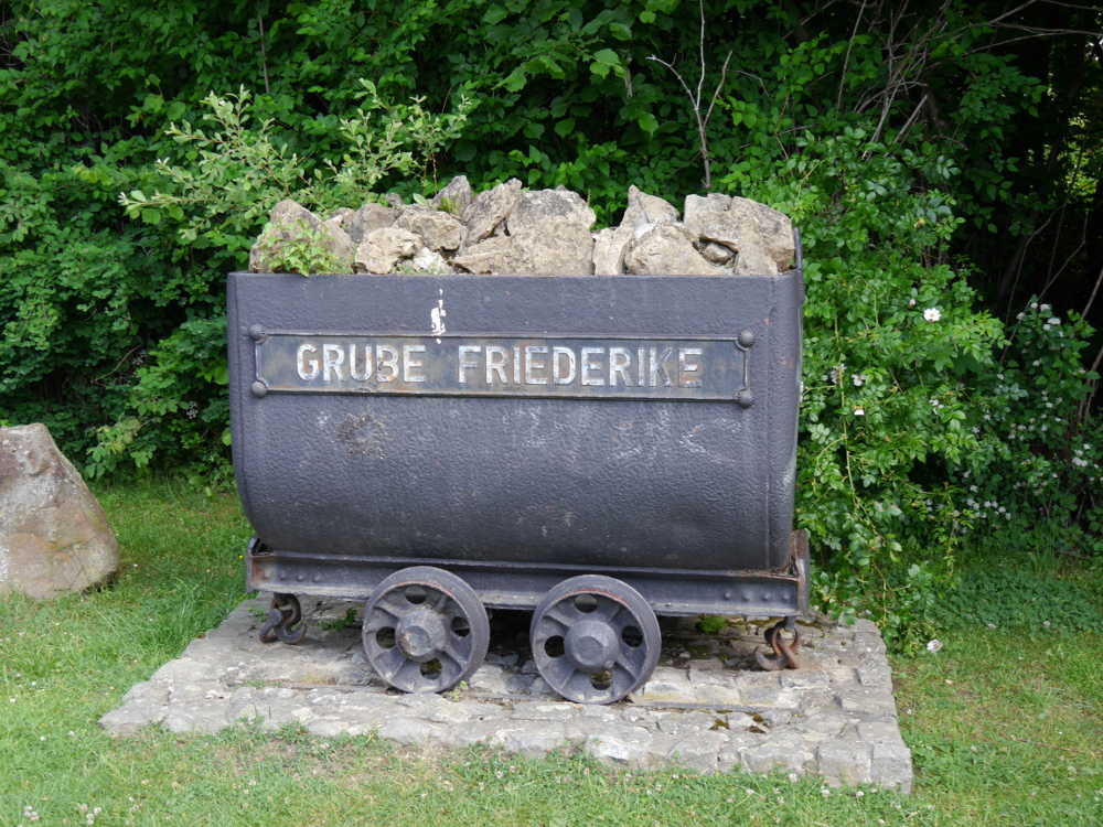 Ein alter Grubenhunt (kleiner Eisenbahnwagen für den Erztranspurt in Bergwerken), gefüllt mit gestein, Beschriftet mit "Grube Friederike" und schon sichtbar in di eJahre gekommen als Denkmal.