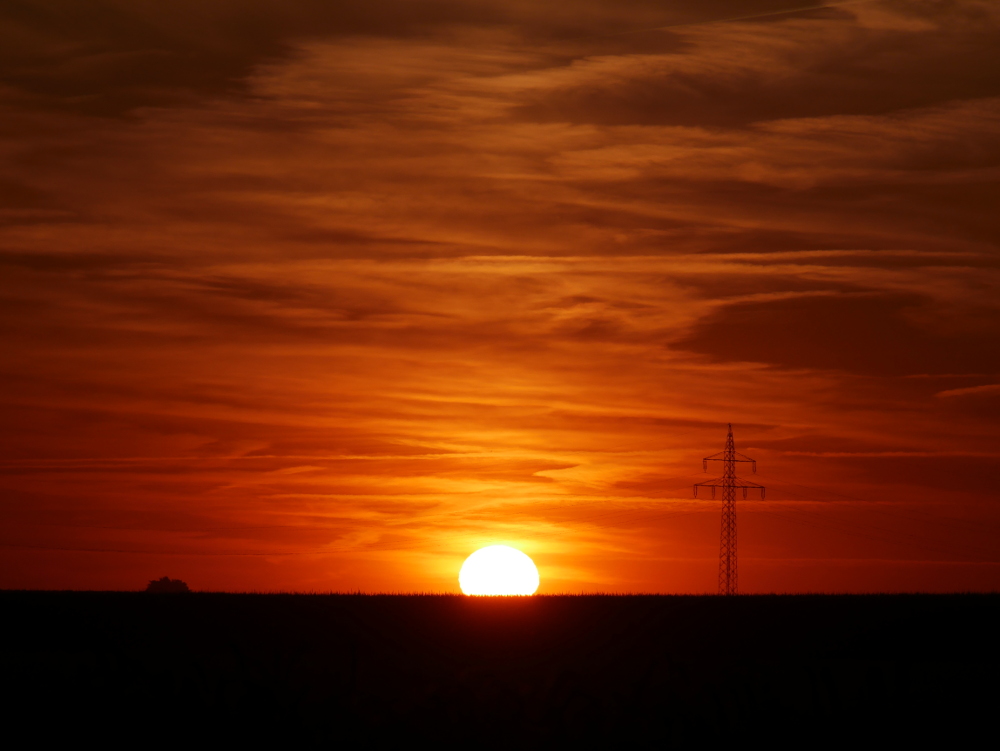 Das Foto zeigt einen dramatischen Sonnenuntergang. Die Sonnenscheibe ist schon zu etwa 1/4 hinter dem Horizont verschwunden, und durch das Sonnenlicht leuchtet der ganze Himmel mit den Wolken in Gelb- und Orangetönen. Rechts neben der Sonne ist ein Freileitungsmast, links neben der Sonne sind in einiger Entfernung aufgestapelte Strohballen zu erkennen.