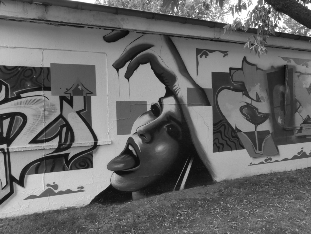 Das s/w-Foto zeigt ein Graffiti-Gemälde auf einer Wand, das zentrale Motiv ist eine Frau mit geöffnetem Mund, die die Zunge rausstreckt. Ihre Hand hält sie über den Kopf und von den Fingern tropft eine Flüssigkeit herab.