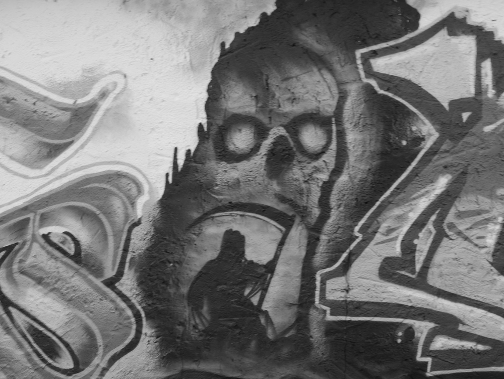Das s/w-Foto zeigt ein Grafiti, das zentrale Motiv ist ein Sensemann mit Sense (als Symbol für den Tod), der dem Betrachter den Rücken zukehrt. Darüber ein Totenkopf, die Sense vom Sensemann ist in etwa der Mund vom Totenkopf.