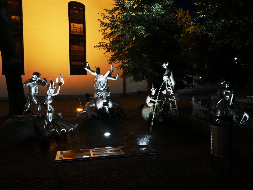 Die Nachtaufnahme von glänzenden Stahlskulpturen, die Sagenfiguren darstellen sollen, welche verschiedene Instrumente spielen. Im Hintergrund erkennt man die weiße Wand einer Kirche