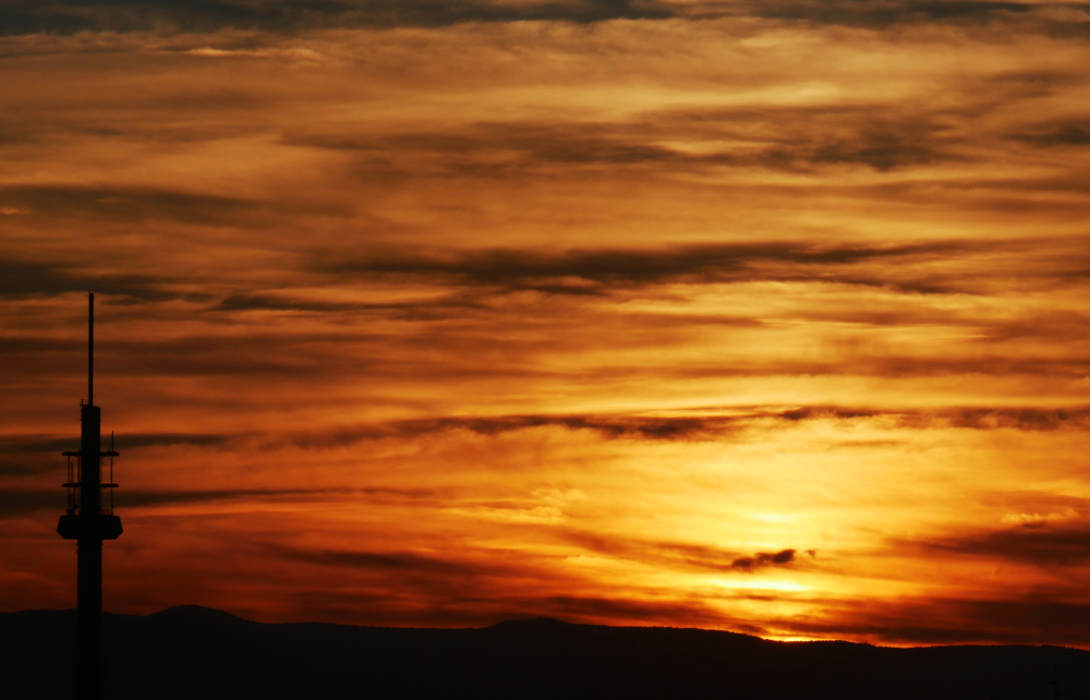 Das Bild zeigt einen dramatischen Sonnenuntergang. Am linken Bildrand ist die Spitze mit den Antennen eines Fernsehturms erkennbar, etwas im rechten Drittel ist hinter Wolken die Sonne. Durch die Wolken und Sonne ist das ganze Bild Orange-Rot.