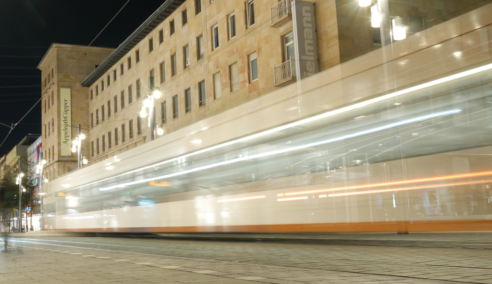 Das Foto zeigt eine Langzeitbelichtung einer Straßenbahn in der Innenstadt, durch die lange Belichtungszeit sind die Lichter usw. der Straßenbahn zu "Spuren" geworden. Die Straßenbahn ist Weiß mit orangenen Streifen.