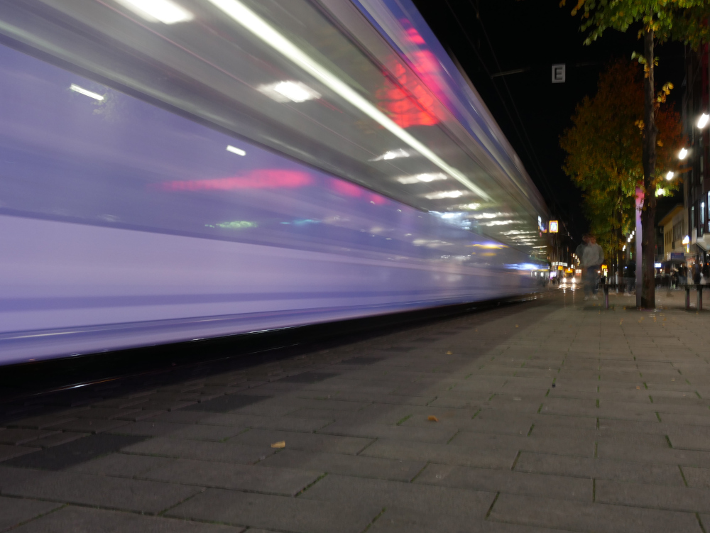 Das Foto zeigt eine Langzeitbelichtung einer Straßenbahn in der Innenstadt, durch die lange Belichtungszeit sind die Lichter usw. der Straßenbahn zu "Spuren" geworden. Die Straßenbahn ist weiß/violett.