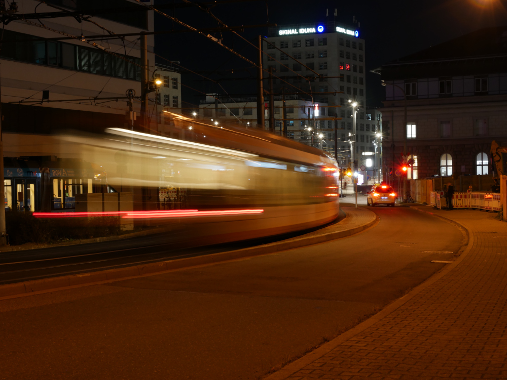 Das Foto zeigt eine Langzeitbelichtung einer Straßenbahn auf Anfahrt zum Mannheimer Hauptbahnhof. Weiter hinten kann man ein Hochhaus mit Signal-Iduna Werbung erkenne, neben der Straßenbahn steht ein Taxi an einer roten Ampel. durch die lange Belichtungszeit sind die Lichter usw. der Straßenbahn zu "Spuren" geworden.