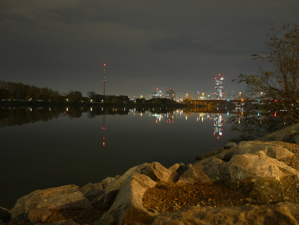Das Foto ist eine nächtliche Langzeitbelichtung in Wien an der Donau. In der flachen Donau spiegeln sich die Lichter, man kann den Fernsehturm, diverse Hochhäuser und auch Steine am Donau-Ufer erkennen. Am rechten Rand ist auch ein wenig Gebüsch erkennbar.