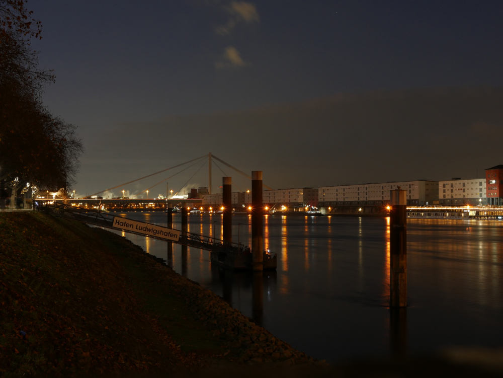 Das Foto zeigt eine nächtliche Landschaftsaufnahme am Rhein zwischen Ludwigshafen und Mannheim. Am linken Ufer ist eine Schiffs-Anlegebücke mit dem Schild "Hafen Ludwigshafen" erkennbar, rechts sind Lagerhäuser und Schiffe erkennbar. Weiter hinten ist die Kurt-Schumacher-Brücke und dahinter sind die Schornsteine usw. des BASF-Werks erkennbar.