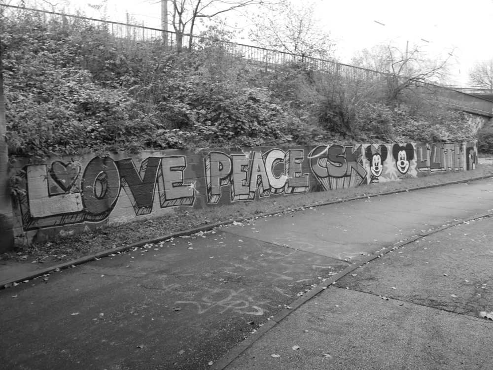 Das s/w-Foto zeigt eine halbhohe Mauer, auf die ein Graffiti gemalt wurde. Das Graffiti zeigt "Love, Peacke, CSK", daneben Mini und Mikey-Maus und daneben "ULith".