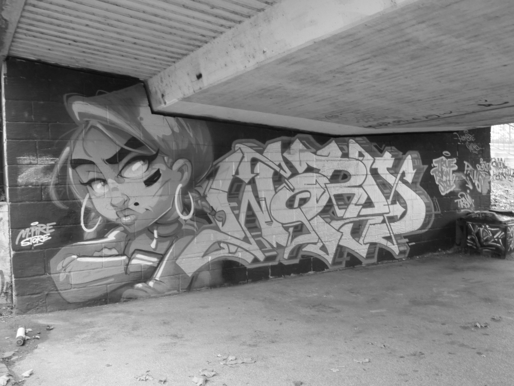 Das s/w-Foto zeigt ein Graffiti auf einem Pfeiler einer Straßenbrücke, man erkennt links Oberkörper und Gesicht einer jungen Frau, deneben ein wildes Graffiti ohne erkennbare Buchstaben.
