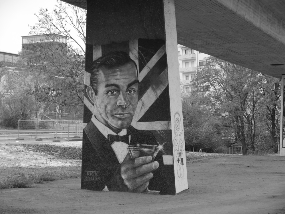 Das s/w-Foto zeigt den Pfeiler einer Straßenbrücke, auf dem Pfeiler ist Gesichts und Oberkörber des fiktiven Geheimagenten "James Bond" als Graffiti vor einer britischen Flagge angebracht. Man sieht die Signatur "Rick Riojas"