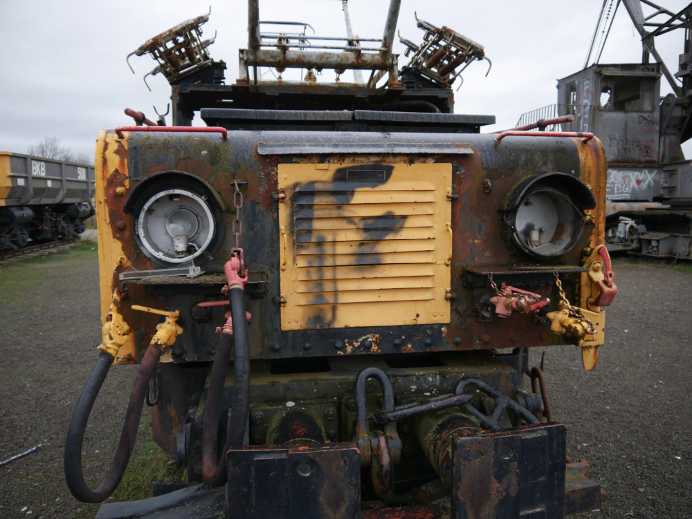 Das Foto zeigt die Spitze einer alten, verrosteten und durch Vandalismus beschädigten Tasgebau-Lokomotive. Im Hintergrund kann man Teile von Wagen und einem Tagebau-Bagger erkennen.