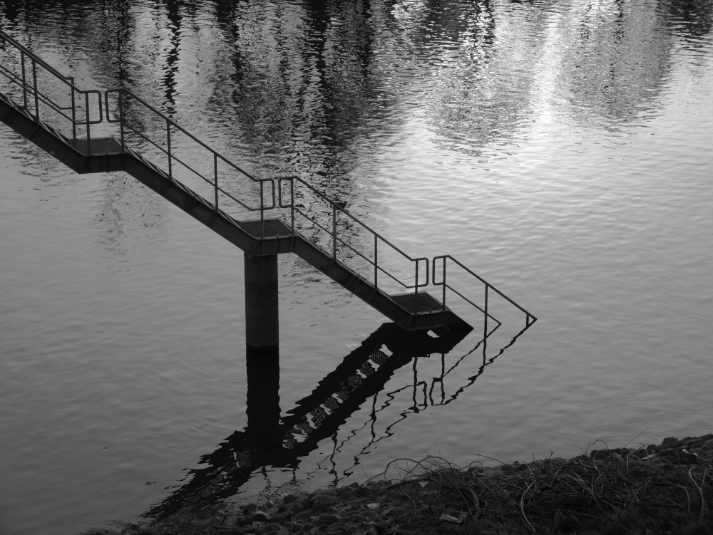 DAs s/w-Foto zeigt eine Treppe (so eine Industrietreppe aus Gitterblech), welche in einem Fluss verschwindet.