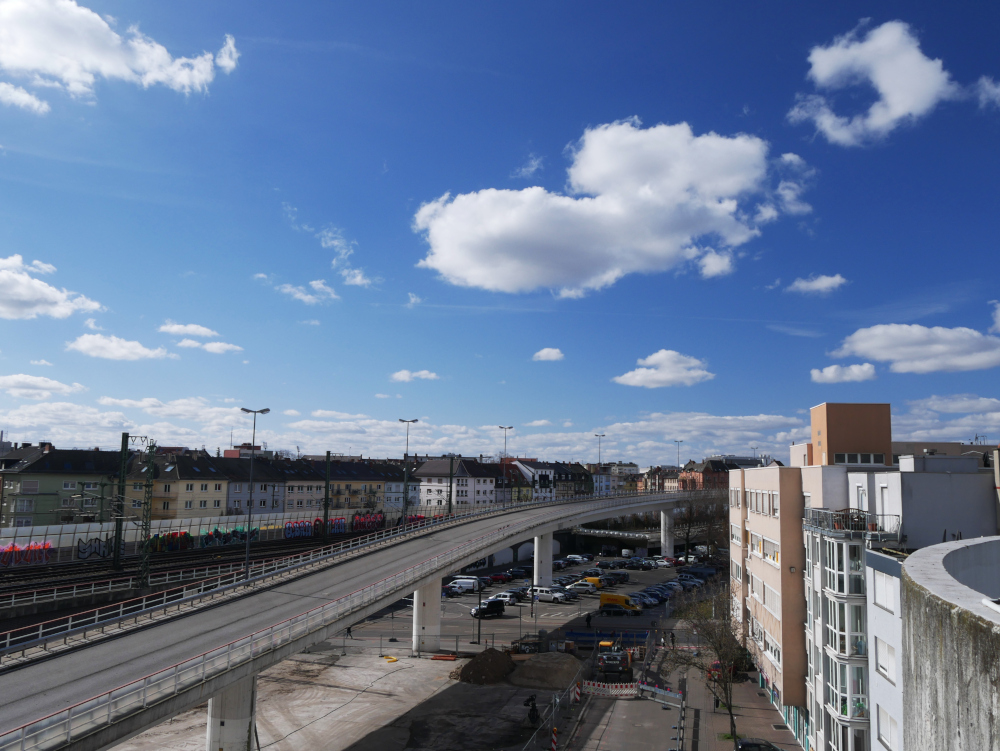 Das Foto zegit eine städtische Landschaftausfnahme. Rechts und Links sind Häuer, dazwischen eine aufgeständerte Hochstraße mit Parkplätzen drunter und eine Bahntrasse. Am blauen Himmel sind viele dramatische weiße Wolken erkennbar.