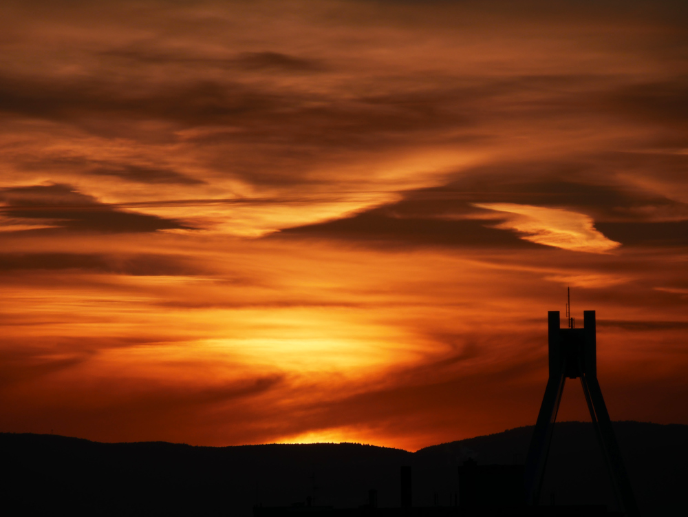 Das Foto zeigt einen Sonnenuntergang. Vor der untergehenden Sonne sind aber Wolkenschichten, die durch die Sonne in nur schwer beschreibbaren Mustern und orange-rot-gelben Farbtönen beleuchtet werden. Rechts sieht man als Silouette die Spitze einer Brücke (Die Straßenbrücke über den Hauptbahnhof von Ludwigshafen)