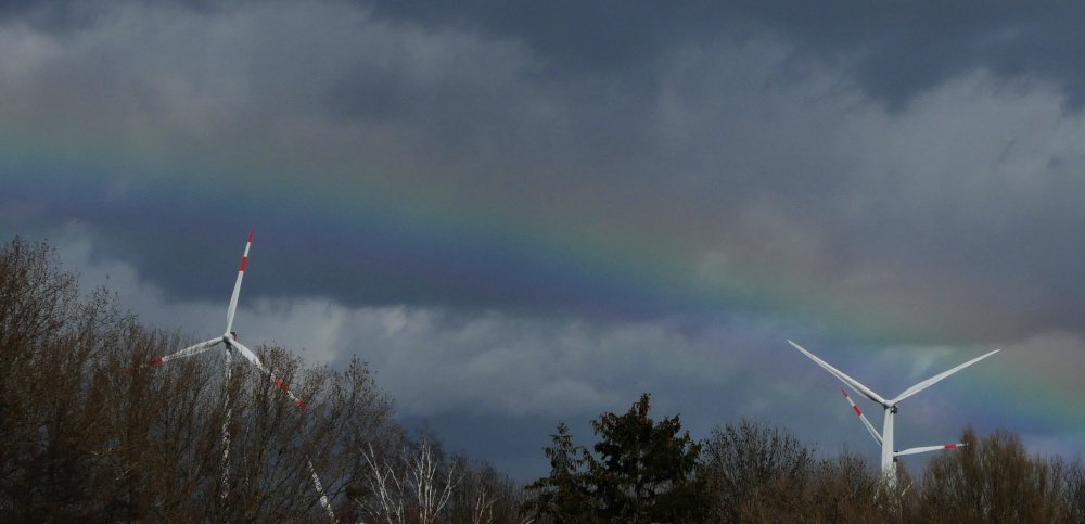 Das Foto zeigt einen Regenbogen vor dichten Wolken. Der Regenbogen ist kurz über bzw. an den Rotoren von Windkraftanlagen, im unteren Bereich des Bildes sind Bäume erkennbar.