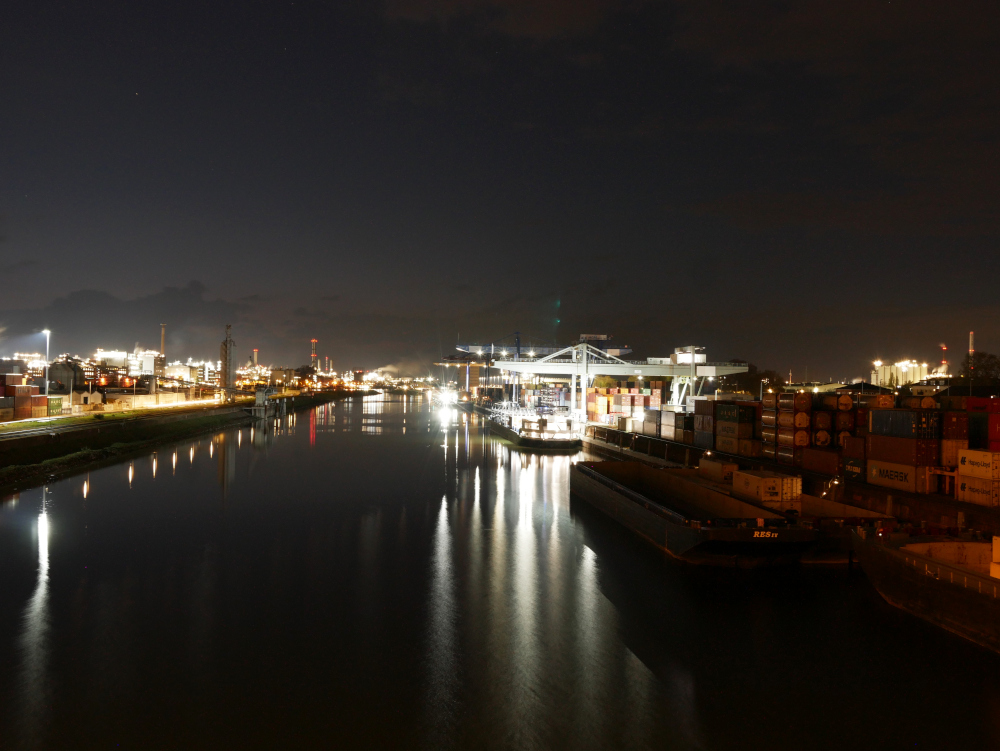 Das Foto zeigt eine nächstliche Landschaftsaufnahme im Hafen von Mannheim. Rechts ist der Container-Kai mit entsprechenden Ladekränen, schiffen und Containerstapeln, dazwischen das Hafenbecken und Links ein beleuchtetes Industriegebiet.