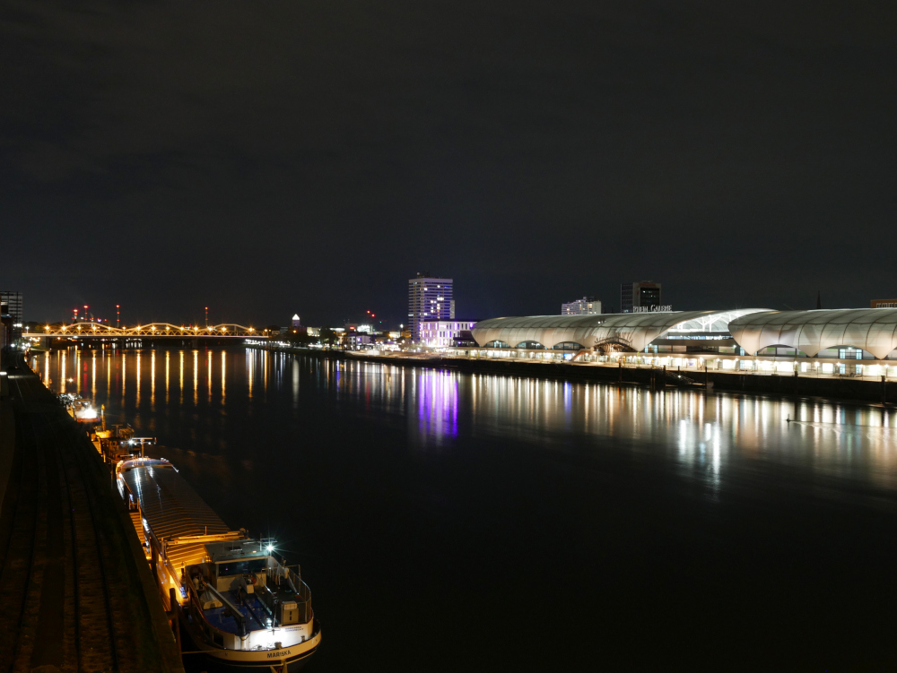 Das Foto zeigt eine Nachtaufnahme am Rhein. Auf der rechten Seite (Ludwigshafen) ist die erleuchtete Rheingalerie (Einkaufszentrum) und dahinter zwei Hochhäuser, auf der rechten (Mannheimer) Seite ein dunkeler Schienenstrang vom Hafengebiet und einige festgemachte Binnenschiffe. Im Hintergrund ist die beleuchtete nächste Rheinbrücke und noch weiter hinten die Schornsteine vom Mannheimer Kraftwerk.