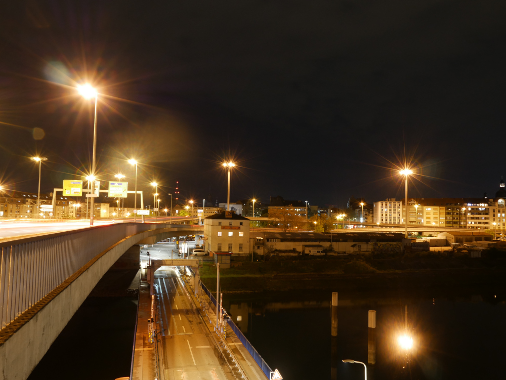 Das Foto zeigt eine Nachtaufnahme an Straßenbrücken im Hafengebiet. Es gibt hier zwei Brücken in gleicher Richtung, aber verschiedener Höhe. Die untere Brücke ist eine zweispurige Klappbrücke über ein Hafenbecken, darüber ist eine mehrspurige Straßen- und Straßenbahnbrücke. Im Hintergrund die Gebäude vom Mannheimer Stadteil Jungbusch.