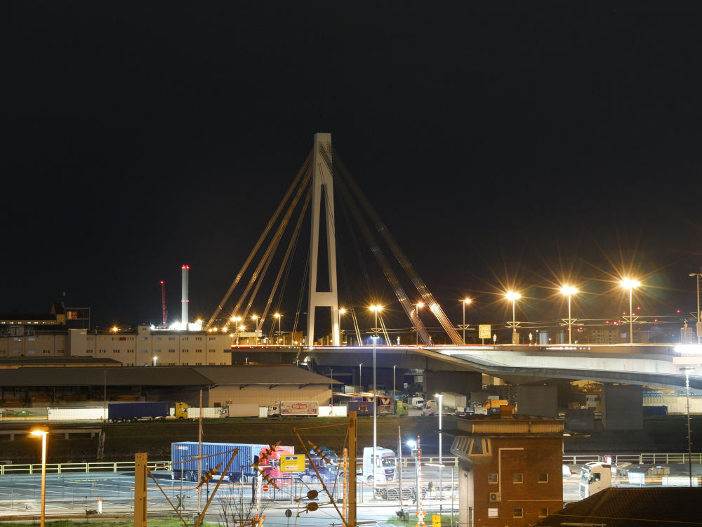 Das Foto zeigt eine Nachtaufnahme der Pylone der Kurt-Schumacher-Brücke zwischen Mannheim und Ludwigshafen. Auf der Brücke kann man auch gut die Oberleitung der Straßenbahn erkennen. Im Vordergrund sind die Gebäude und ien paar LKW des Mannheimer Hafenviertels.