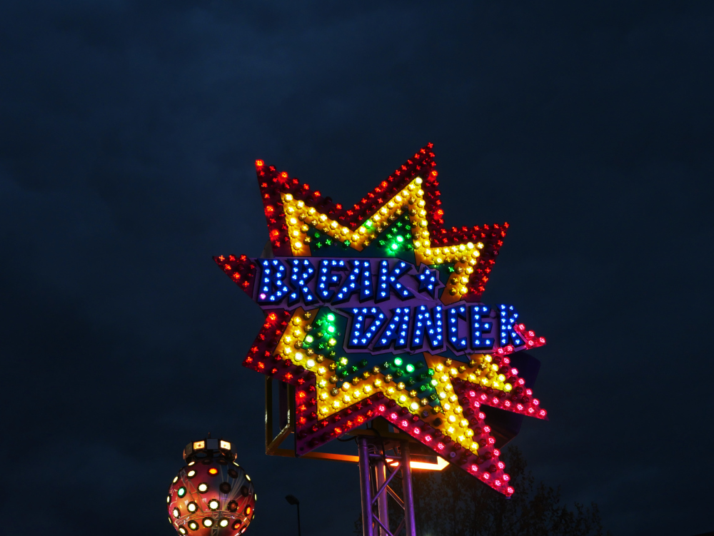 Das Foto zeigt ein großes, durch bunte Lichter gebildetes Schild in Sternform mit der Aufschrift "Break Dancer". Es gehört erkennbar zu einem Schausteller-Betrieb. Danhinger sieht man Teile einer Leuchtkugel und darüber schwarzen Himmel.