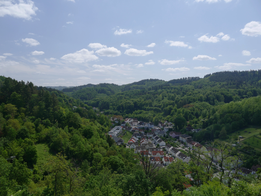 Das Foto zeigt eine Landschaftsaufnahme, im Tal sind einige Häuser, daneben sind grün bewaldete Berghänge und darüber blauer Himmel mit weißen Wolken. Die Aufnahme ist erkennbar auf einem der Berge gemacht und zeigt den Blick nach unten ins Tal