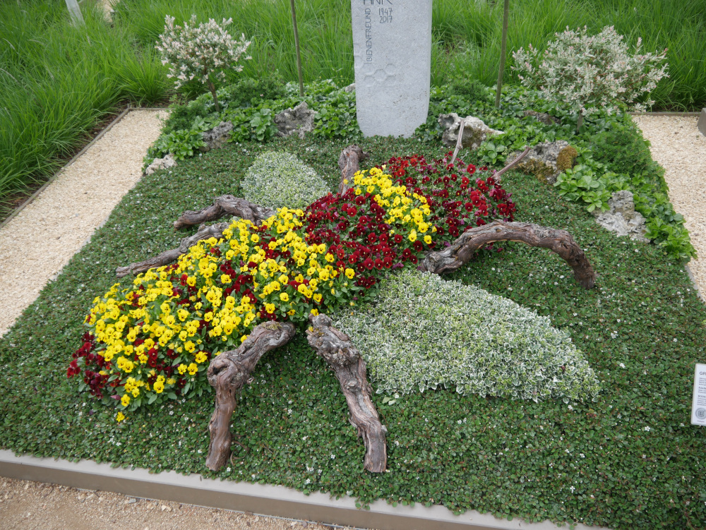 Eine künstlerische Grabgestaltung aus Ästen, Blumen und Bodendeckern, die zusammen die Form einer Hommel ergeben. Im Hintergrund die Grabsteele.