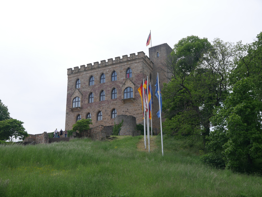 Das Foto zeigt das Hambacher Schloß bei Neustadt/Weinstraße. Auf dem Schloß weht die Deutsche Fahne, vor dem Schloß sind Fahnen masten mit weiteren Fahnen, die aber nicht alle erkennbar sind.