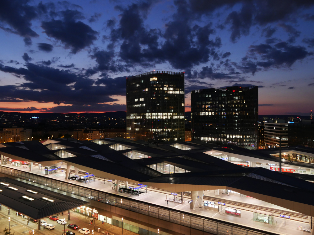 Das Foto zeigt eine Nachtaufname vom Hauptbahnhof in Wien. Das Foto ist aus erhöhter Position in einem Hochhaus aufgenommen und Zeigt die Bahnsteiganlagen von oben, dahinter sind Hochhäuser zu sehen. Darüber ist der schon sehr dunkele Himmel mit ein paar Wolken, im Hintergrund ist noch ein klein wenig Restglühen vom Sonnenuntergang erkennbar.