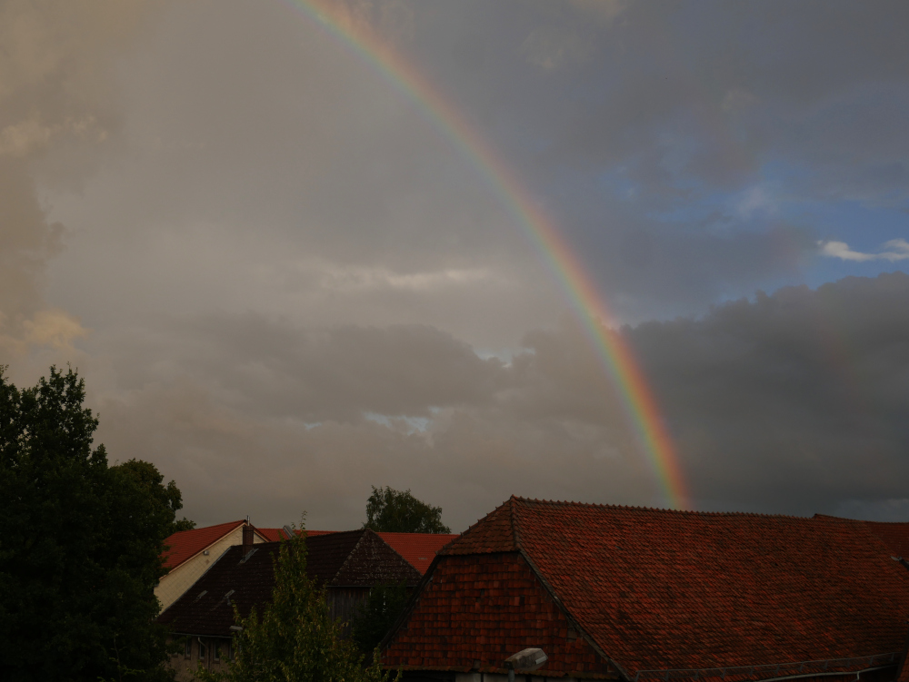 Das Foto zeigt einen Regenbogen über den Dächern eines Dorfs. Zwischen den Dächern sieht man auch hohe Bäume.