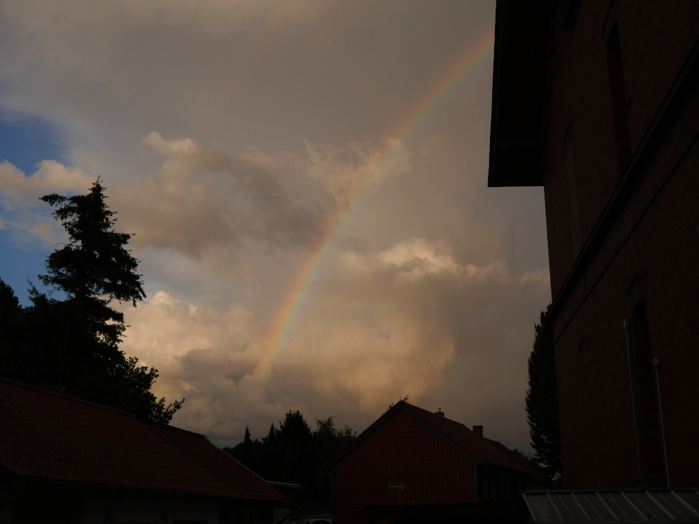Man sieht einen Regenbogen über einem Dorf. Rechts ist ein relativ hohes Backsteinhaus, welches das Bild am ganzebn Rand dominiert und zum linken Rand hin ist dann der wolkige Himmel mit dem Regenbogen zu sehen.