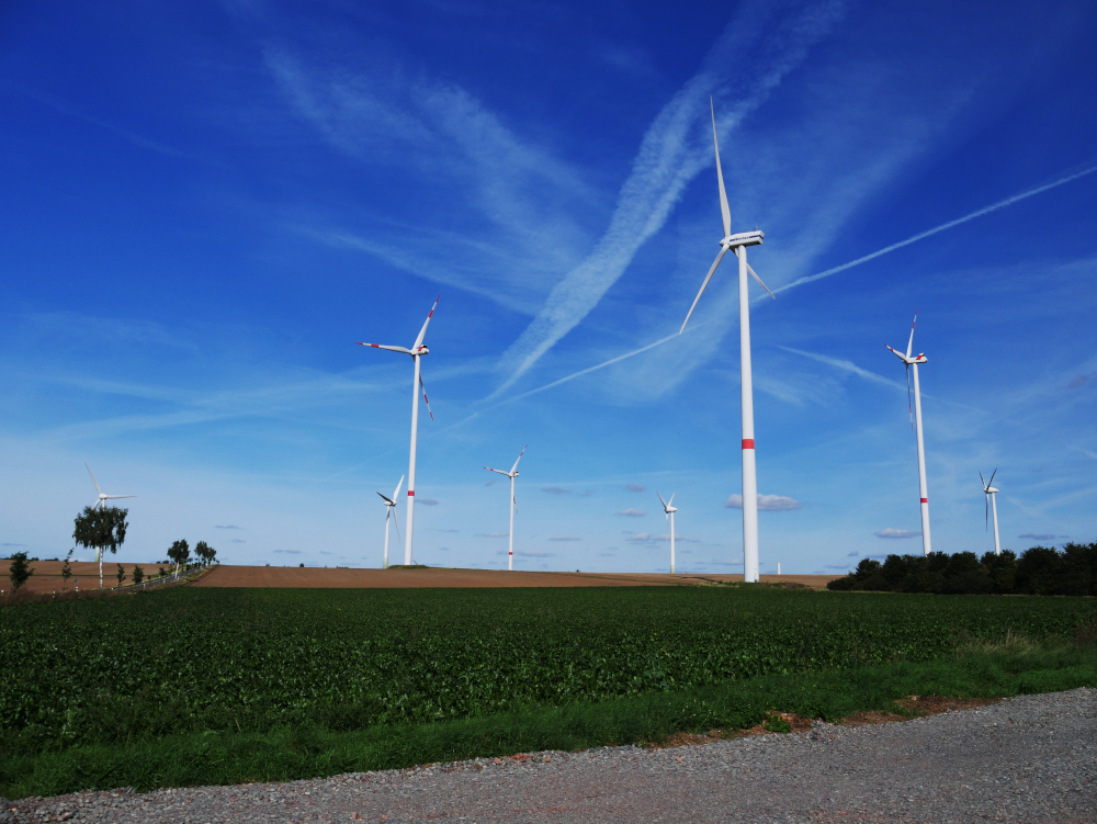 Ds Foto zeigt Windkraftanlagen unter blauem Himmel, dazwischen sind Felder, Bäume/Büsche und am linken Bildrand läuft eine Straße durch.