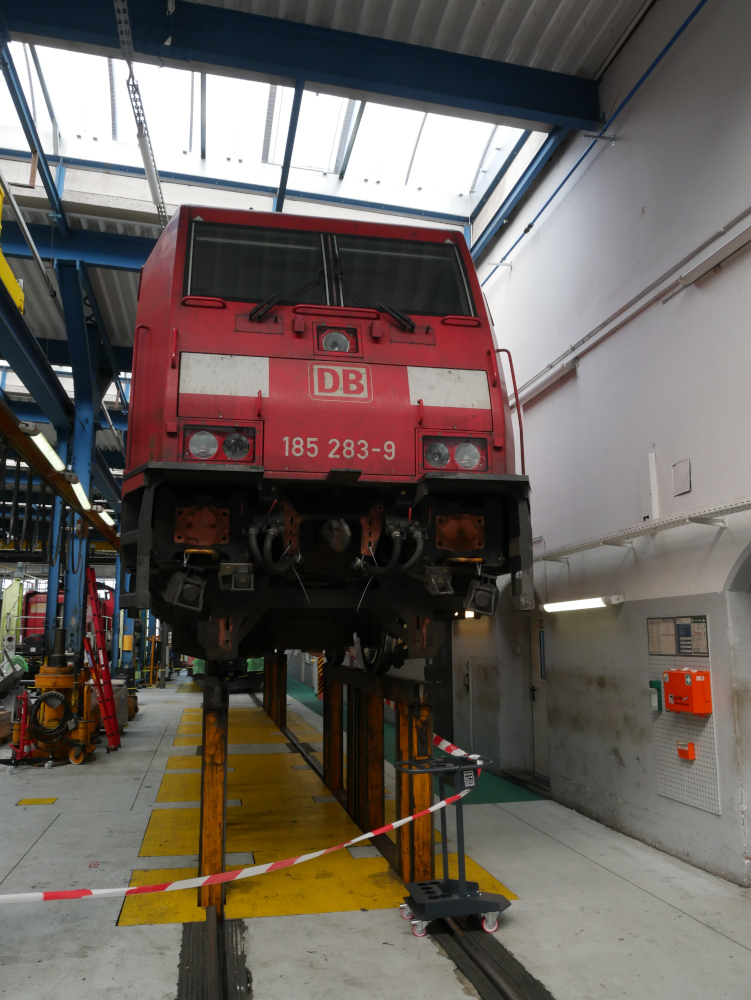 Das Foto zeigt die rote Elektrolok 185 283-9. Diese ist derzeit auf einer Art Hebebühne für Züge ca 1,5m in die Höhe gehoben. Die vorderen Puffer und Kupplungsteile sind teilweise zur Reparatur demontiert.