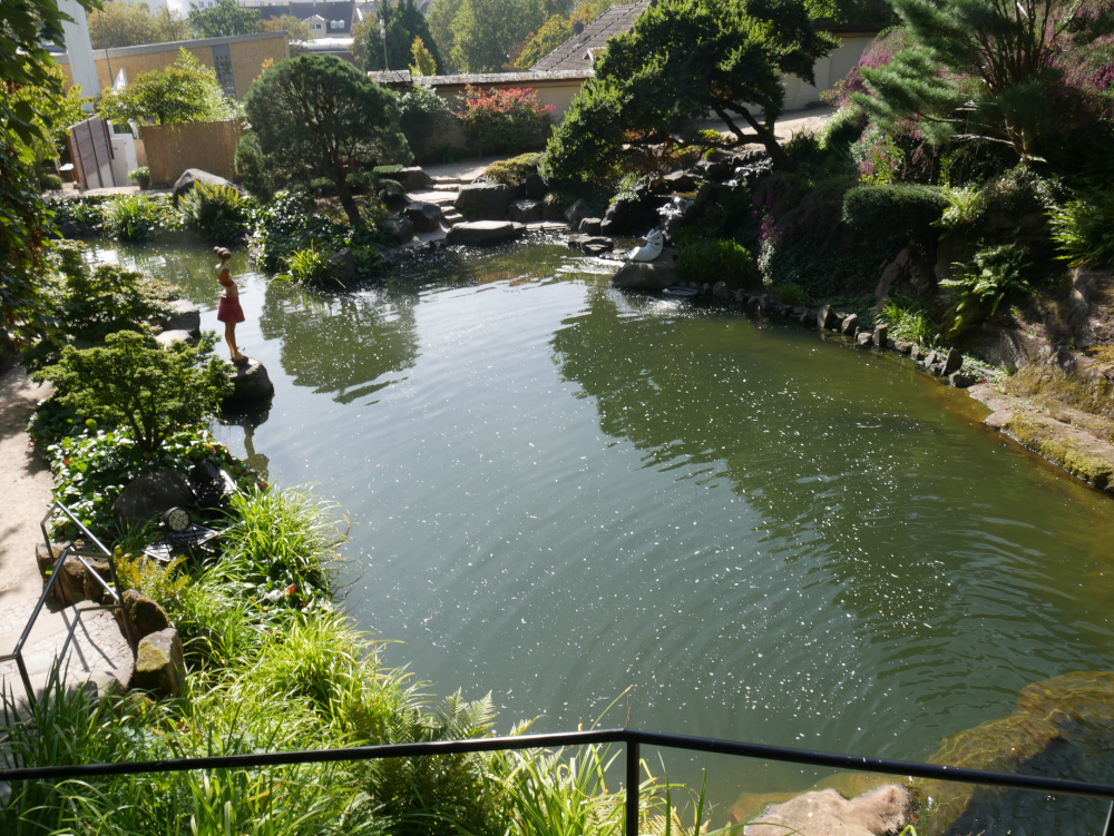 Eine Landschaftssaufnahme im japanischen Garten in Kaiserslautern. Man sieht einen Tech, drum herum Pflanzen, Steine und Wege. Am linken Ufer steht eine weibliche, menschengroße Skulptur.