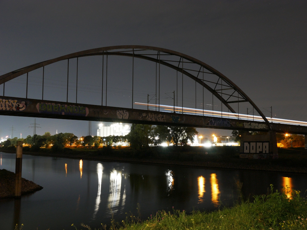 Das Foto zeigt eine mit Graffiti beschmierte Bogenbrücke für die Eisenbahn über den Neckar in Mannheim. Es ist eine Langzeitbelichtung, dadurch ist der Zug zu einem Lichtstreifen geworden, der von rechts kommend bis Etwa in die Mitte der Brücke gefahren ist. Im Hintergrund hinter der Brücke ist ein beleuchtetes Industriegebiet erkennbar.