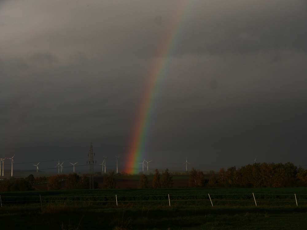Die Landschaftsaufnahme zeigt einen Regenbogen, der im Bereich eines Windparks beginnt und nach oben weg geht. Bis zum Windpark sieht man Felder, eine Baumreihe und eine Hochspannungs-Freileitung, der Himmel über dem Horizont ist mit grauen Wolken verhangen, vor denen sich der Regenbogen schön abhebt.