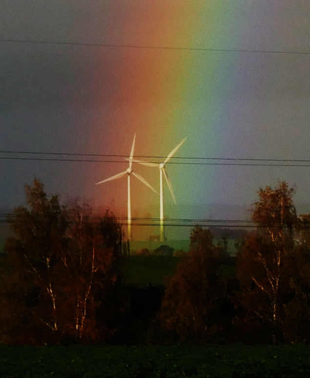 Ds Foto zeigt zwei Windkraftanlagen, welche im bunten Streifen eines Regenbogens stehen.