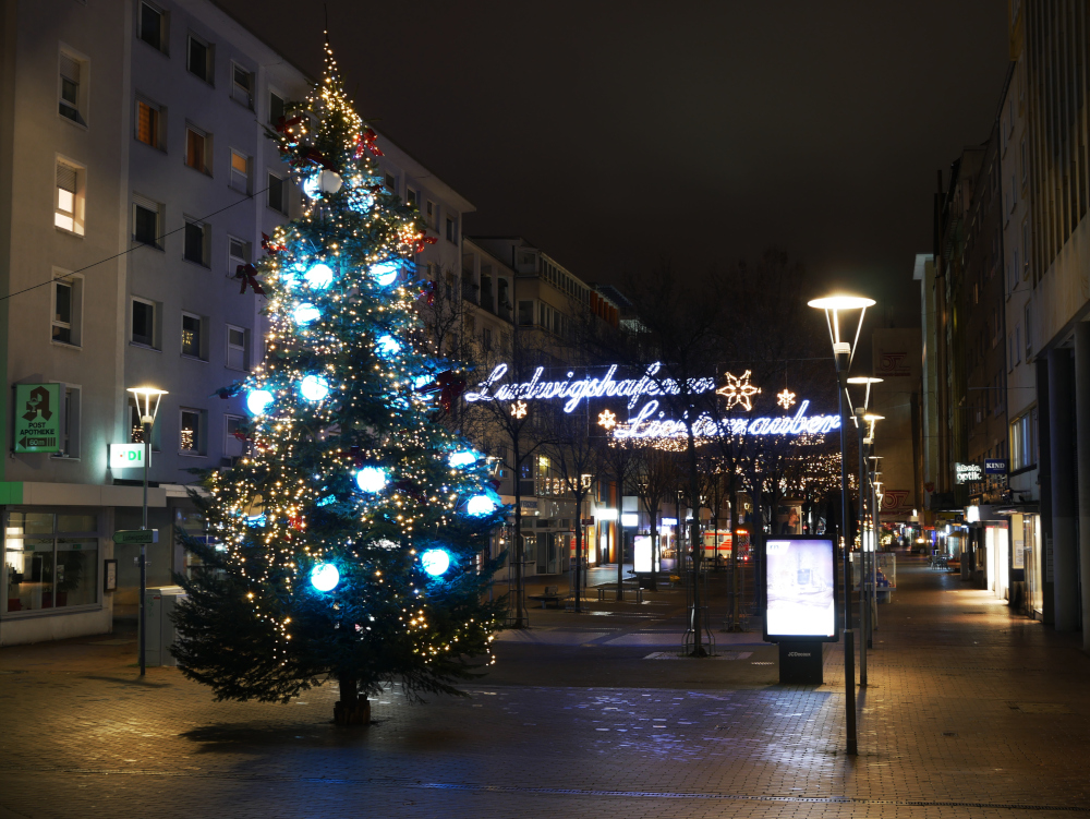 Das Foto zeigt die Weihnachts-Lichtinstallation in der Innenstadt von Ludwigshafen (Fußgängerzone). Vorne steht ein Weihnachtsbäume mit großen, beleuchteten Kugeln, danach ist auf Höhe der Laternen der Leuchtschriftzug "Ludwigshafener Lichterzauber" gespannt, dahinter sieht mann immer wieder Lichternetze über der Straße.