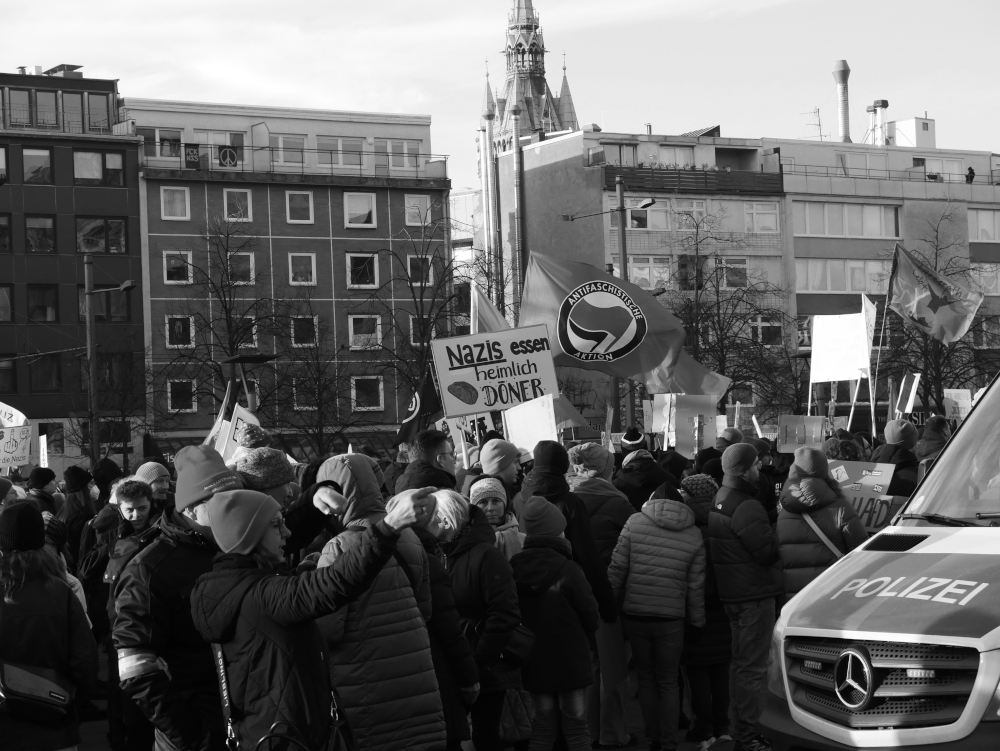 Das s/w-Foto wurde bei einer Demo gegen Rechtsextremismus am 20.01.2024 in Braunschweig aufgenommen. In der Bildmitte hält jemand ein Schild "Nazis essen heimlich Döner" hoch, dahinter eine Antifa-Flagge. Rechts das Vorderteil von einem Kleinbus der Polizei, dazwischen Demonstranten.