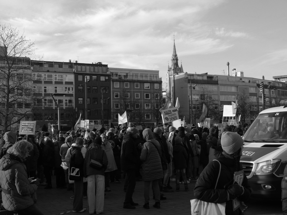 Das s/w-Foto zeigt eine Demo gegen Rechtsextremismus im Braunschweig am 20.01.2024. Man kann rechts das Vorderteil von einem Kleinbus der Polizei erkennen, leicht rechts neben der Mitte hält jemand ein Plakat mit der Aufschrift "Nazis essen heimlich Däönder hoch". Die weiteren Plakate sind schwer erkennbar.