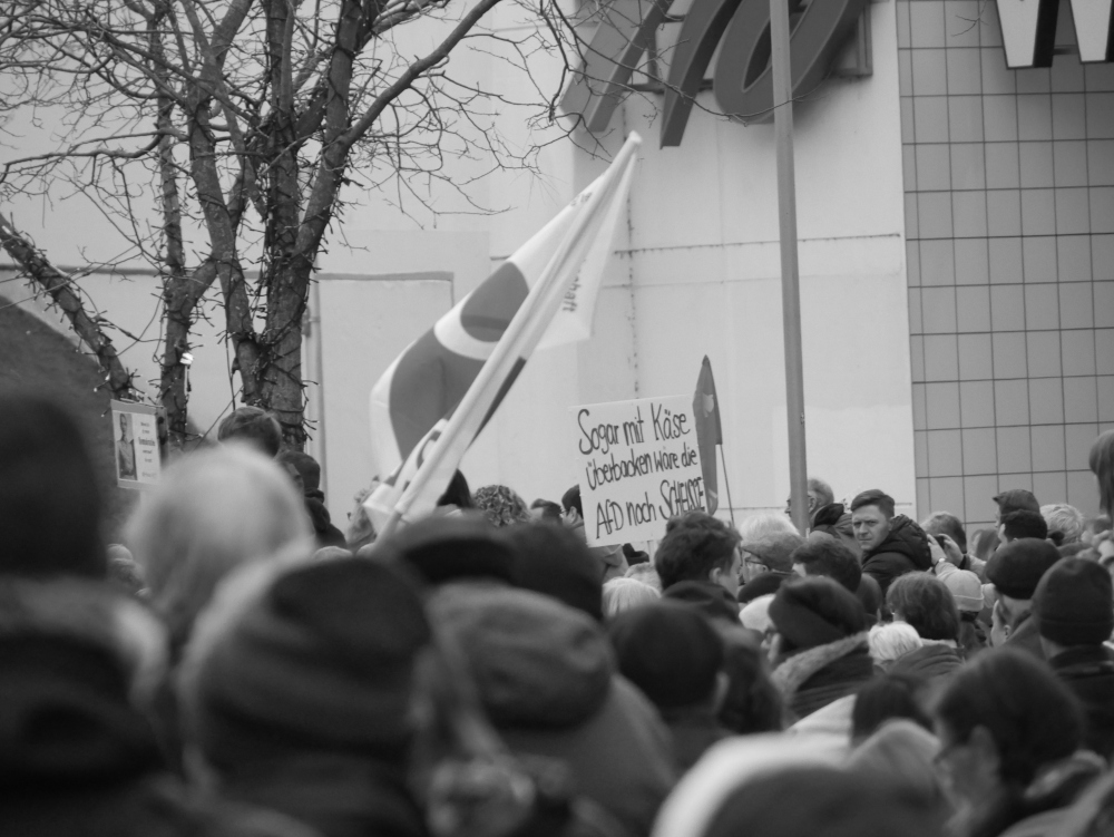 Das s/w-Foto wurde bei einer Demo gegen Rechtsextremismus und Fremdenhass am 03.02.2024 in Ludwigshafen aufgenommen. Man sieht über den Köpfen der Demonstranten ein schild mit dem Slogan "Sogar mit Käse überbacken wäre die AfD n och Scheisse". Daneben Fahnen.