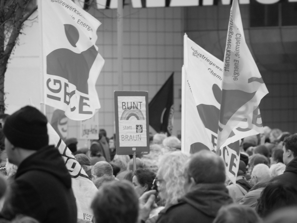 Das s/w-Foto wurde bei einer Demo gegen Rechtsextremismus und Fremdenhass am 03.02.2024 in Ludwigshafen aufgenommen. Man sieht ein Schild "Bunt statt Braun" zwischen IGBCE- und anderen Fahnen