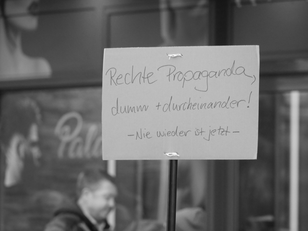 Das s/w-Foto wurde bei einer Demo gegen Rechtsextremismus und Fremdenhass am 03.02.2024 in Ludwigshafen aufgenommen. Es zeigt ein Pappschild mit dem Slogan" Rechte Propaganda, dumm + durcheinander ! Nie wieder ist jetzt"