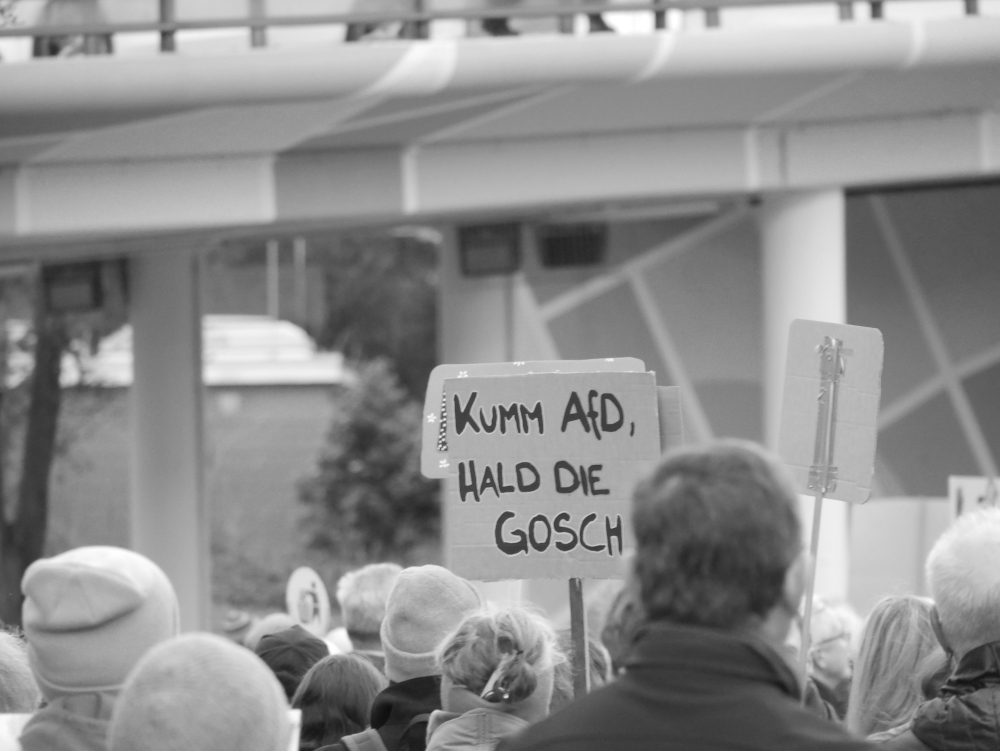 Das s/w-Foto wurde bei einer Demo gegen Rechtsextremismus und Fremdenhass am 03.02.2024 in Ludwigshafen aufgenommen. Man sieht ein Pappschild mit der Aufschrift in Pfälzer Mundart "Kumm AfD, Hald die Gosch".