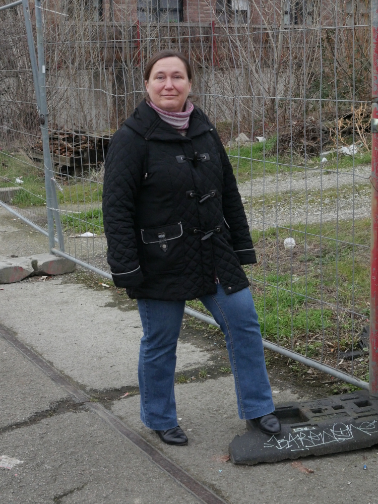 Eine Frau steht in einer schwarzen Jacke und blauer Jeans an einem Bauzaun und hat ihren rechten Fuß auf eines der Bodengewichte für den Zaun gestellt