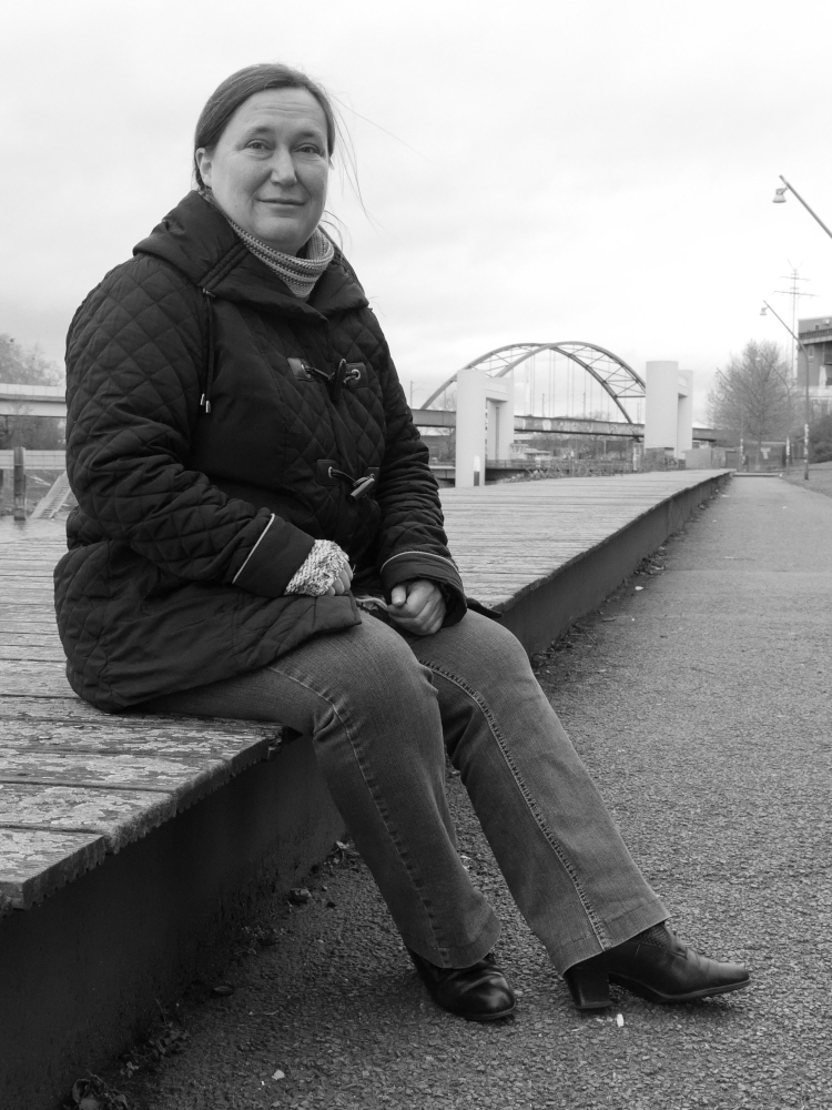 Das s/w-Foto zeigt eine Frau in schwarzer Jacke und blauer Hose die auf einem Holzsteg sitzt. Im Hintergrund ist eine Hubbrücke und eine Bogenförmige Eisenbahnbrücke erkennbar