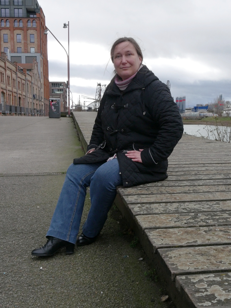 Das s/w-Foto zeigt eine Frau in schwarzer Jacke und blauer Hose die auf einem Holzsteg sitzt. Im Hintergrund sind Industriegebäude, teilweise auch sehr alte, erkennbar.