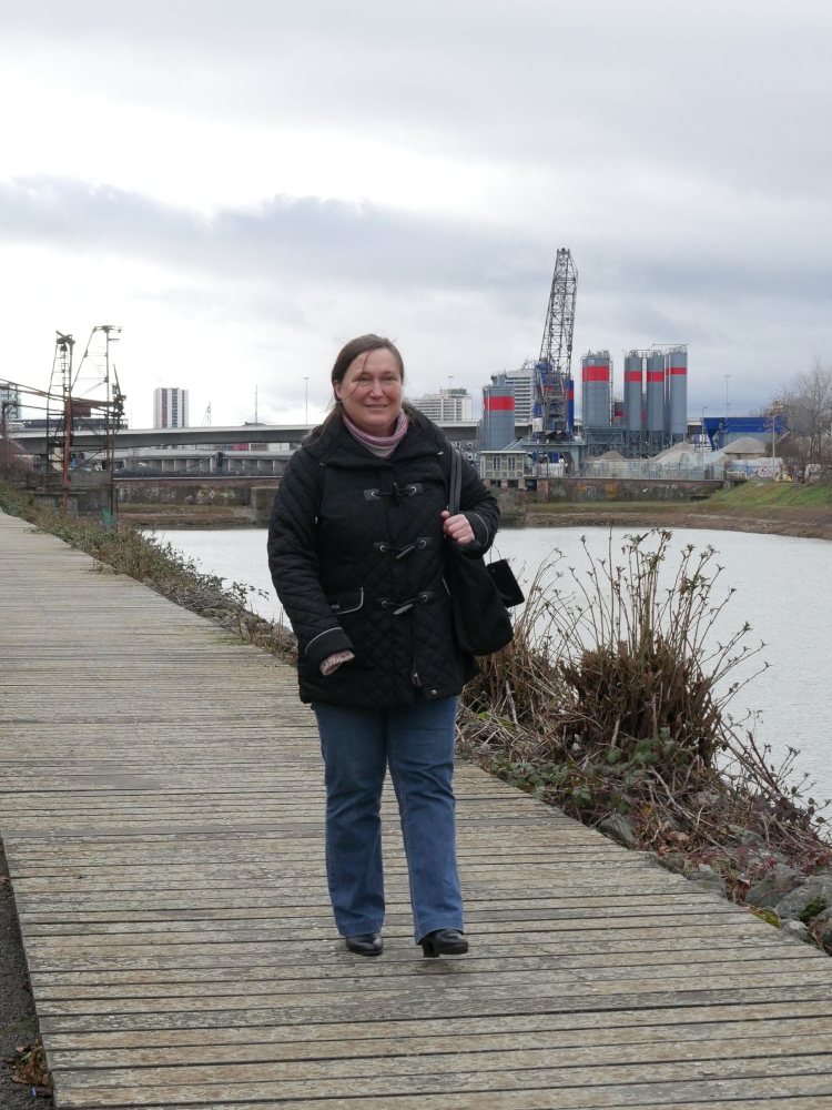 Eine Frau geht über einen Holzsteg am Verbindungskanal in Mannheim, im Hintergrund ist ein Industriegebiet, Straßen und noch weiter hinten Hochhäuser erkennbar. Die Frau trägt eine schwarze Jacke und eine Blaue Jeans.