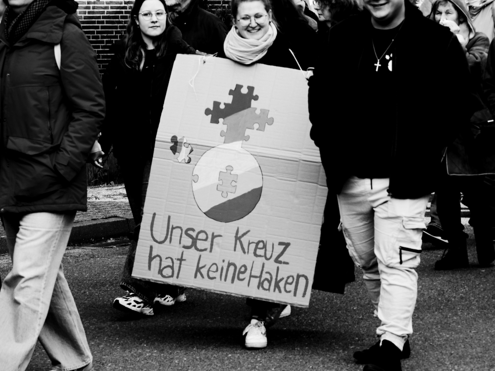 Das s/w-Foto stammt von einer Demo für Demokratie und gegen Rechtsextremismus. Man sieht Teilnehmende, eine trägt ein Papp-Plakat mit dem Logo der Ev. Jugend der Propstei Schöppenstedt und dem Zusatz "Unser Kreuz hat keine Haken"